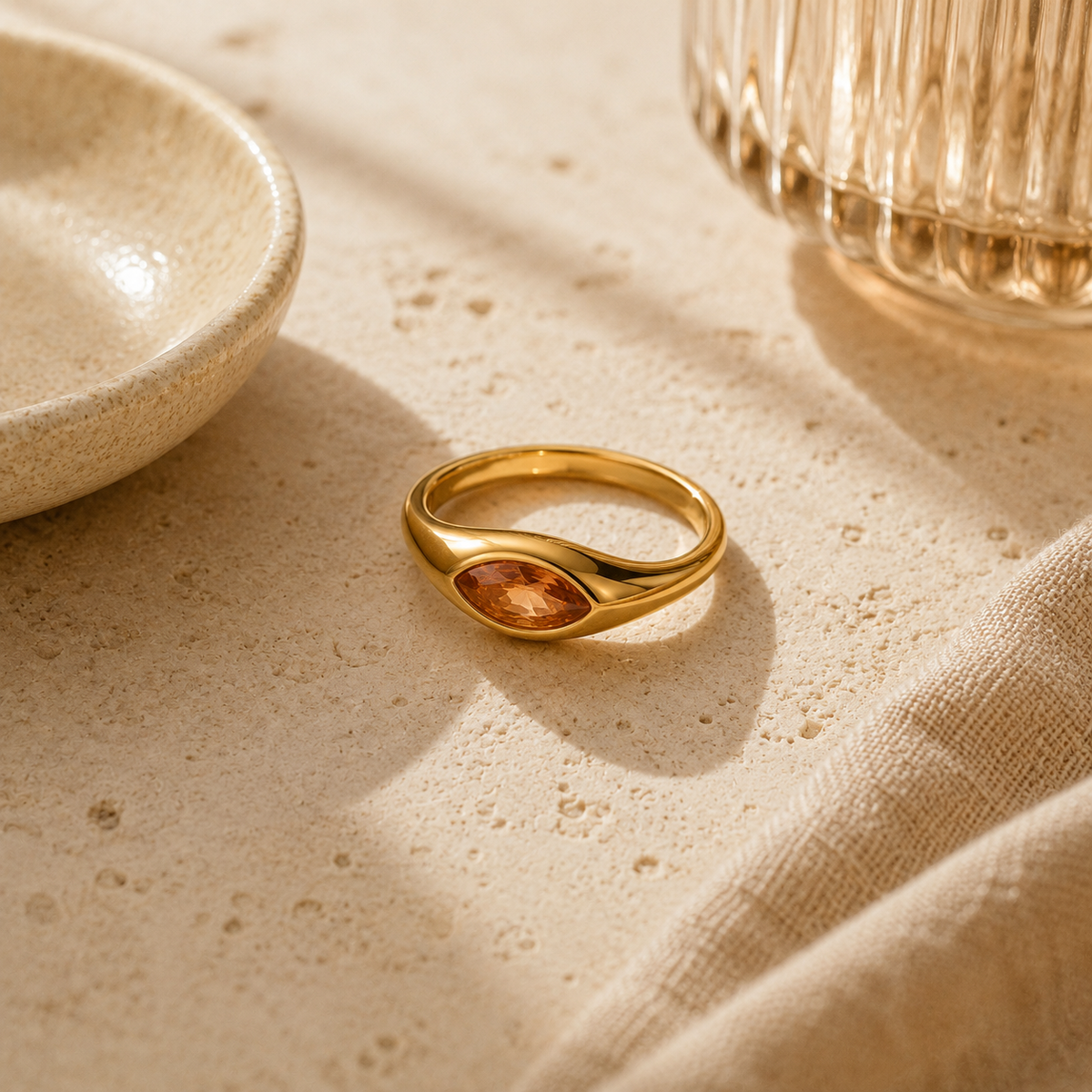 Elegant gold ring with oval amber gemstone, set on textured stone surface with ceramic bowl and glass